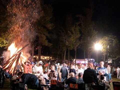 A tall bonfire burning at night with guests in chairs gathered around it