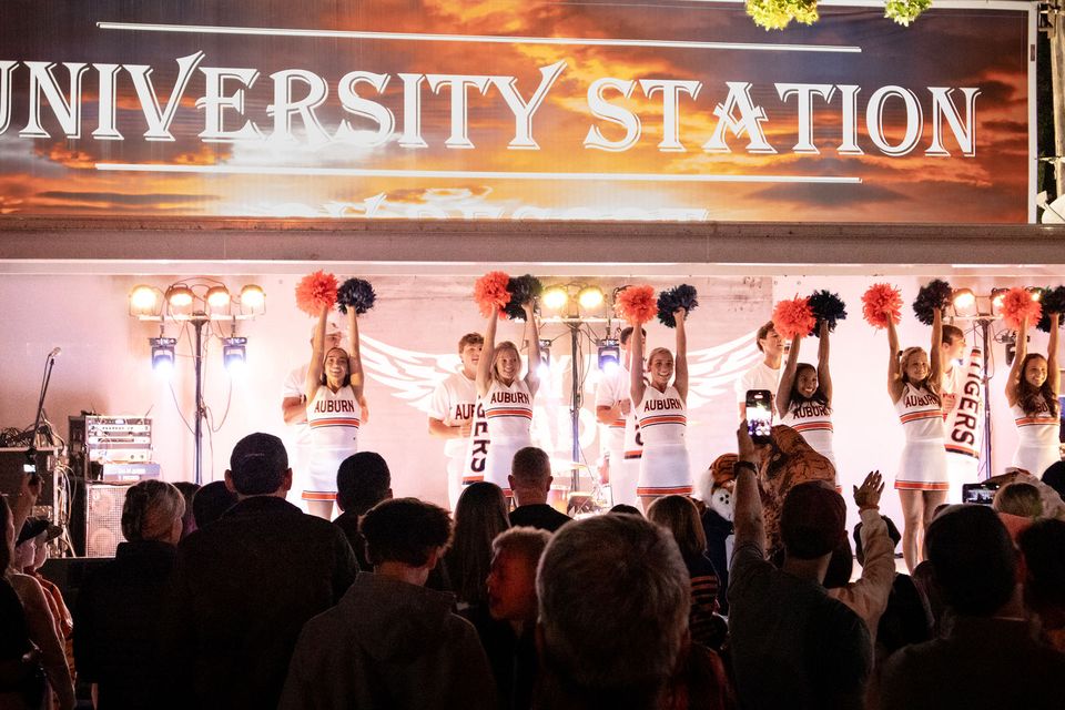 Auburn cheerleaders on the University Station stage at night, pom-poms raised, with the crowd in front of them